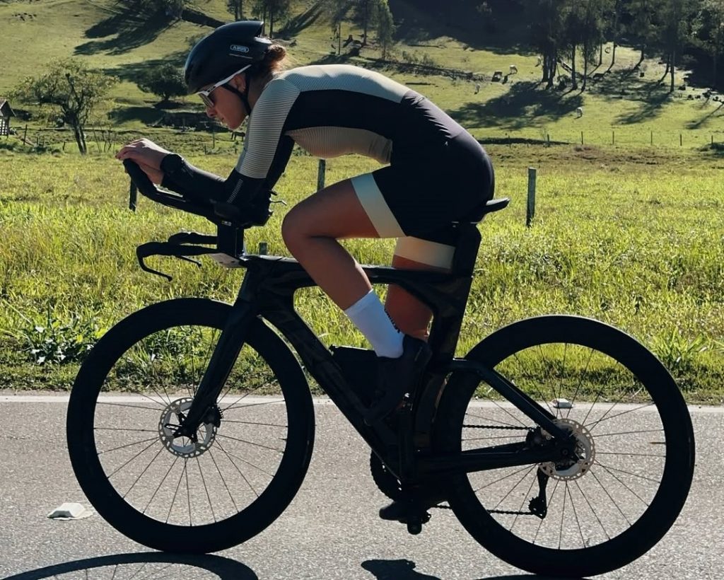 Julia Quarezemin andando de bike Mulher, branca, jovem, andando de bicicleta, fazendo um treino para uma prova de triatlon. Veste Macacão preto com detalhes amarelos e apropriado para a prática. Ela está andando de bicicleta em uma área rural