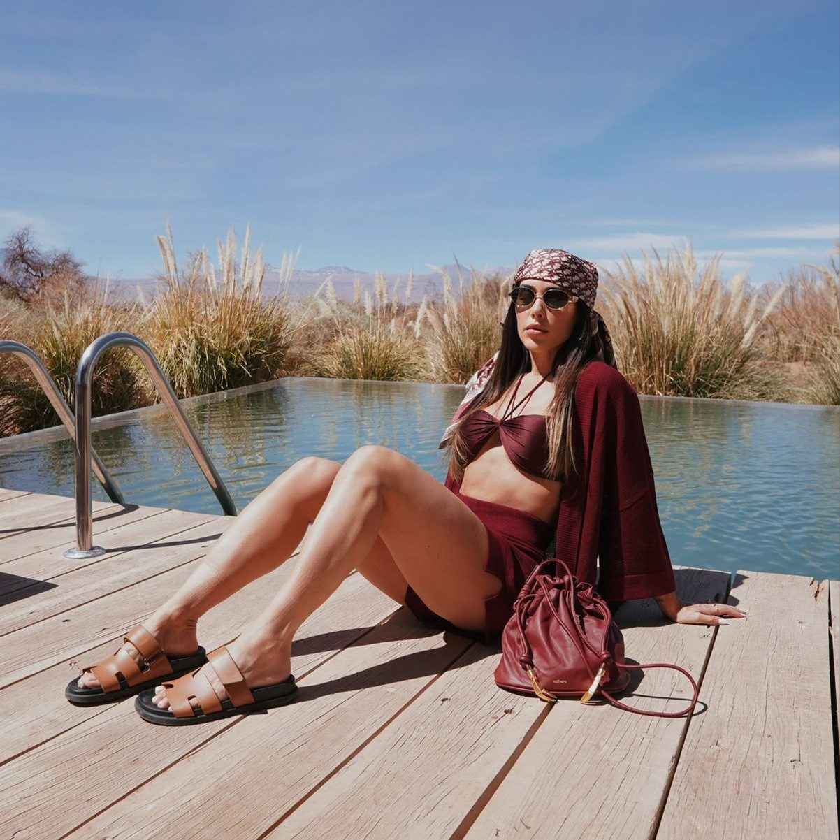 Bella Falconi, uma mulher morena, sentanda em um deck, com vista para o deserto do atacama. Veste um conjunto de saida de praia feito em tricô na cor bordô.