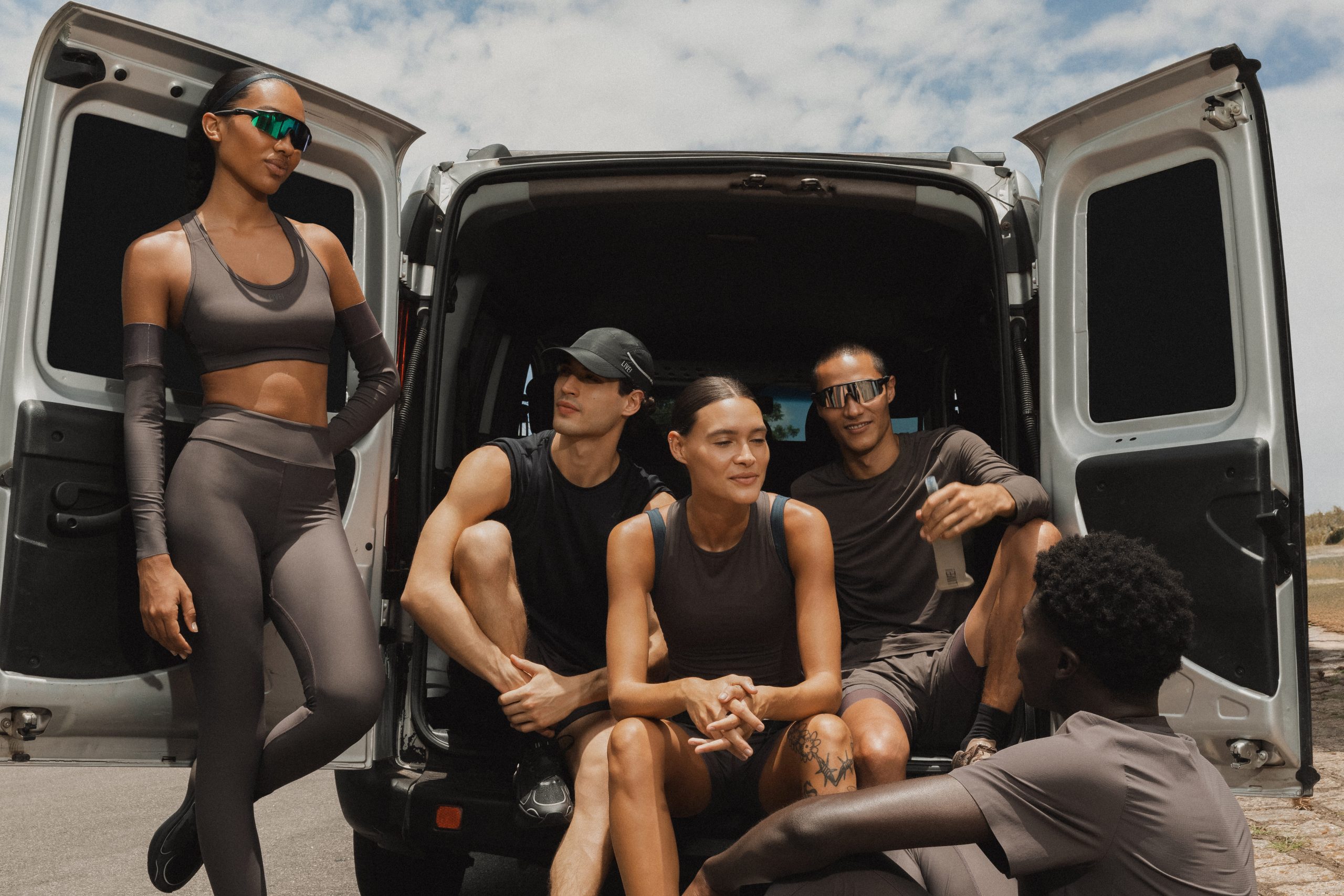 Fotografia horizontal ao ar livre mostra cinco pessoas em roupas esportivas Grafeno Fresh Pro reunidas na traseira de uma van com portas abertas. Uma mulher está em pé à esquerda, enquanto três pessoas sentam no porta-malas e outra interage à direita.